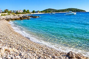 A pebble beach with clear blue water, boats, and buildings in the background. - Apartment Zita (Rogoznica)