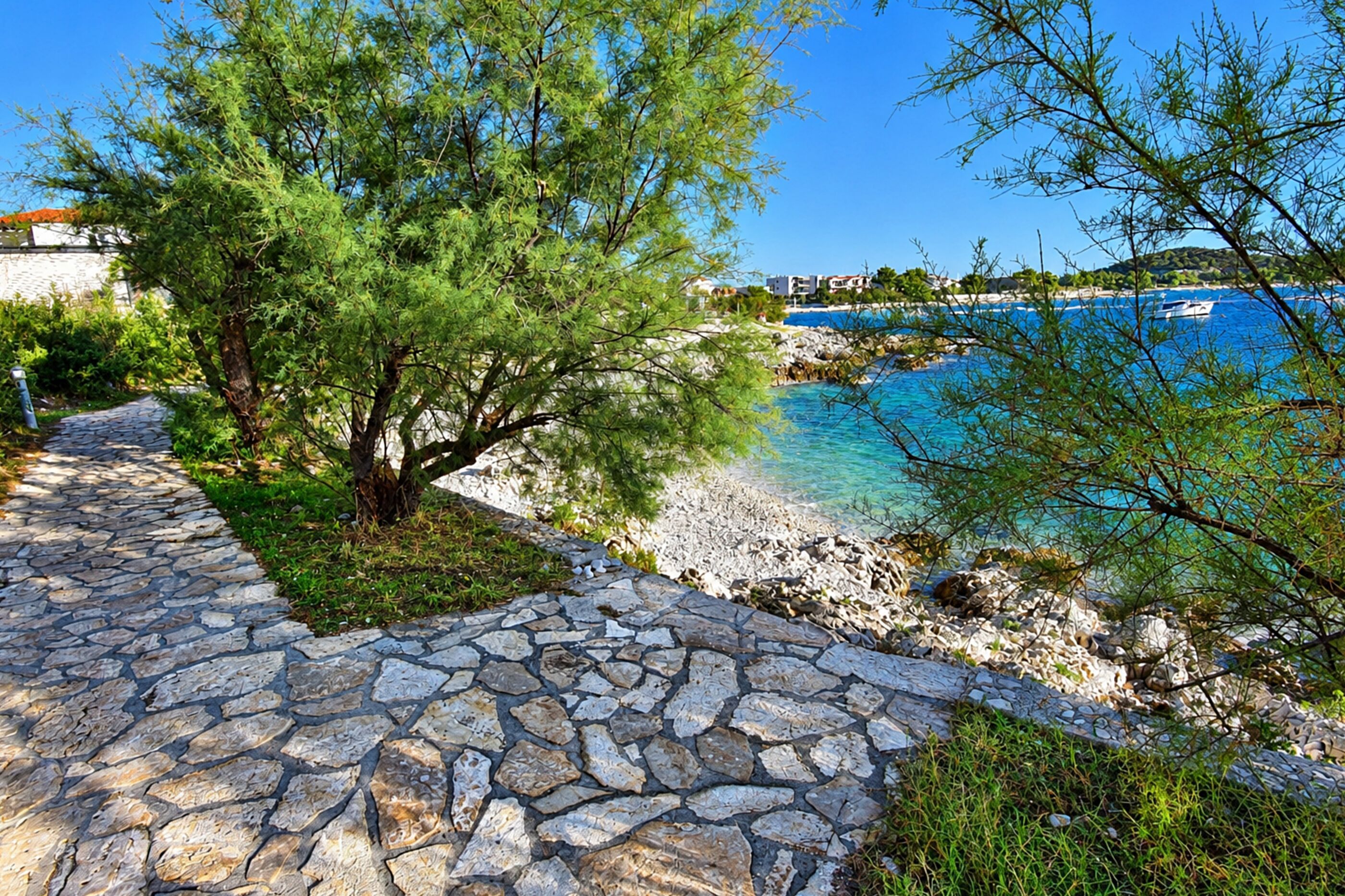 A stone pathway leading to a beach with clear blue water and boats.