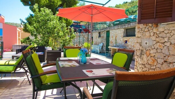 An outdoor patio with a table set for dining, surrounded by green chairs, and a red umbrella providing shade. - Apartments Roko (Primosten)