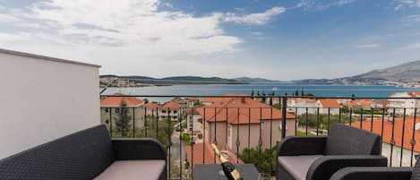 A balcony with wicker furniture, a bottle of champagne, and a glass. The view includes a body of water and buildings in the distance.