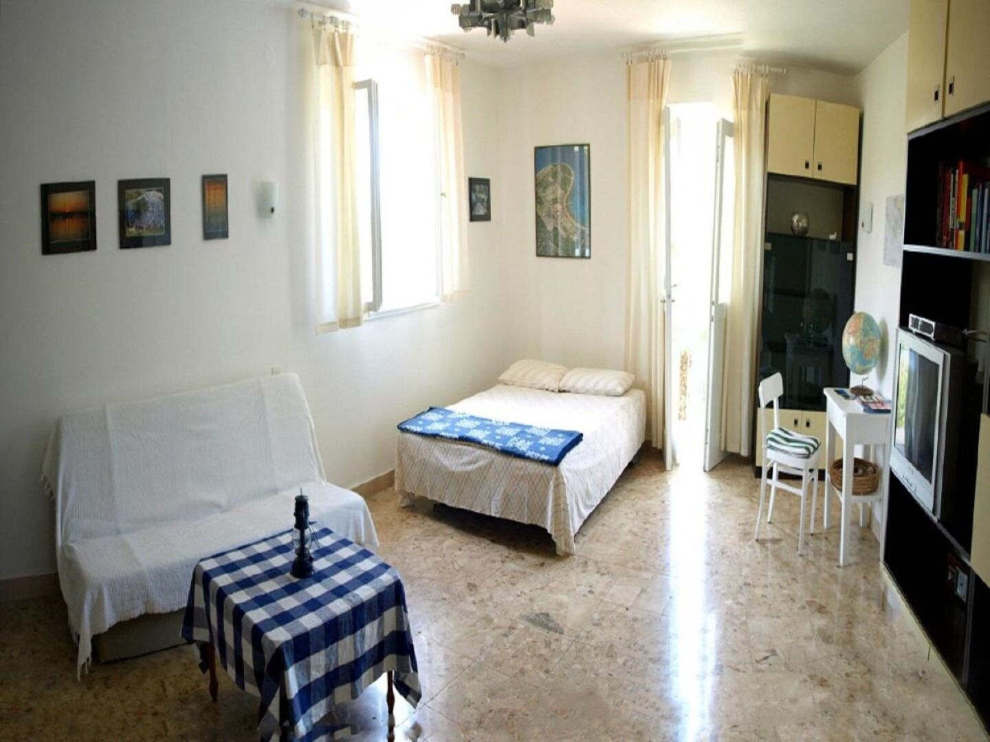 A bedroom with a bed, a small table with a checkered tablecloth, a chair, a bookshelf, and a globe.