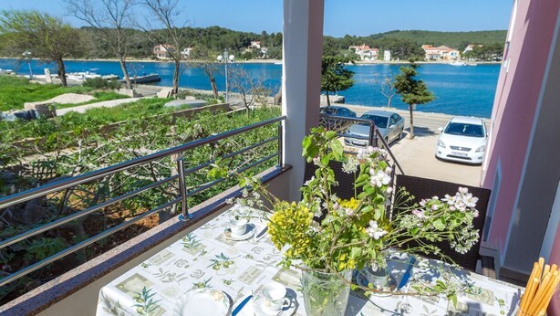 Un balcon avec une table dressée pour le dîner, offrant une vue sur une étendue d’eau et un village côtier.