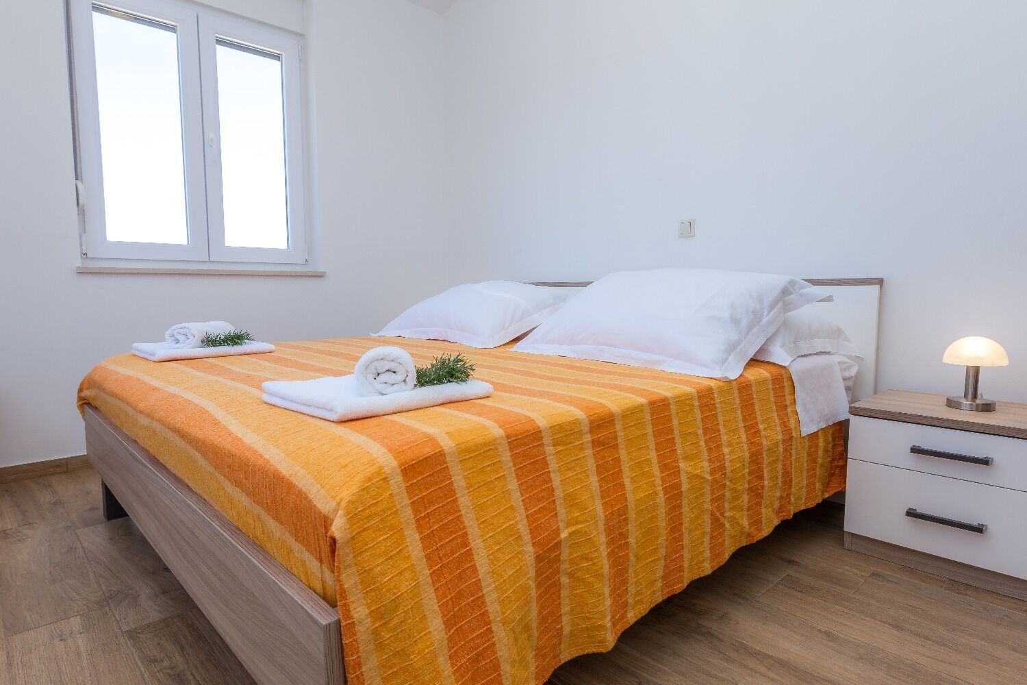Una cama bien hecha con colcha a rayas naranja y amarilla, almohadas blancas y toallas enrolladas sobre un piso de madera.