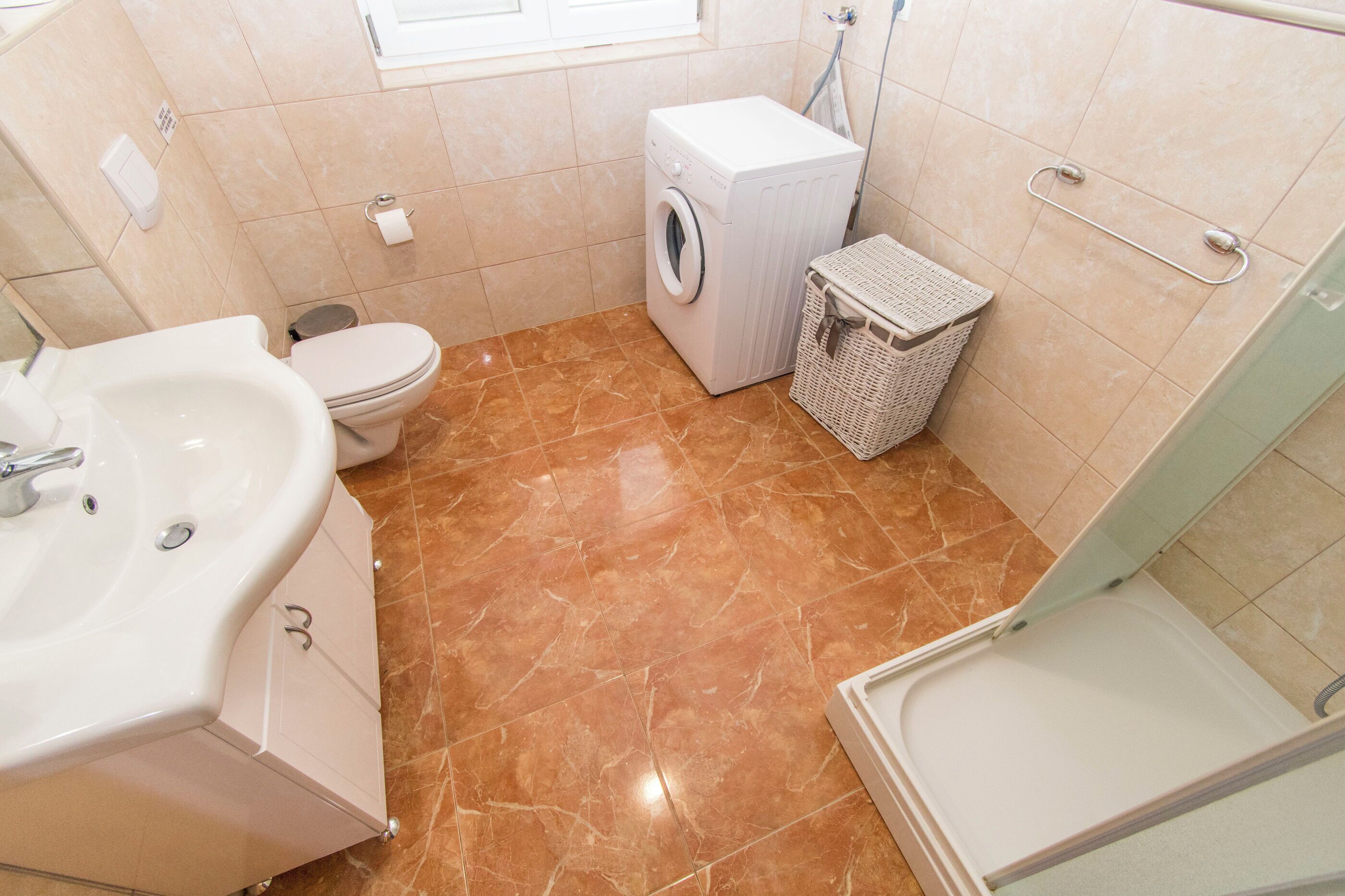 A compact bathroom with a sink, toilet, washing machine, and a small basket.