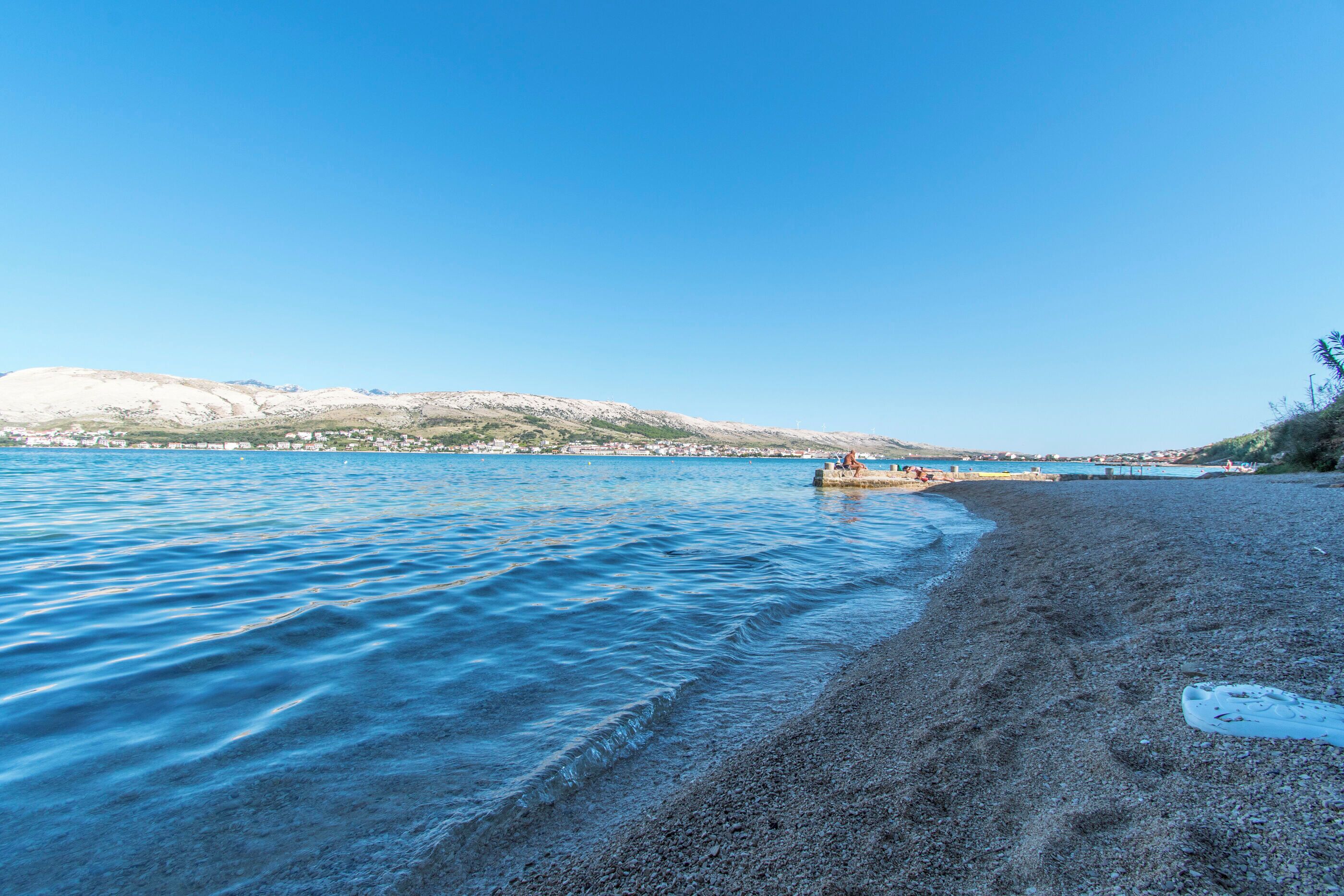 Una playa con aguas azules cristalinas, orilla pedregosa y colinas a lo lejos.
