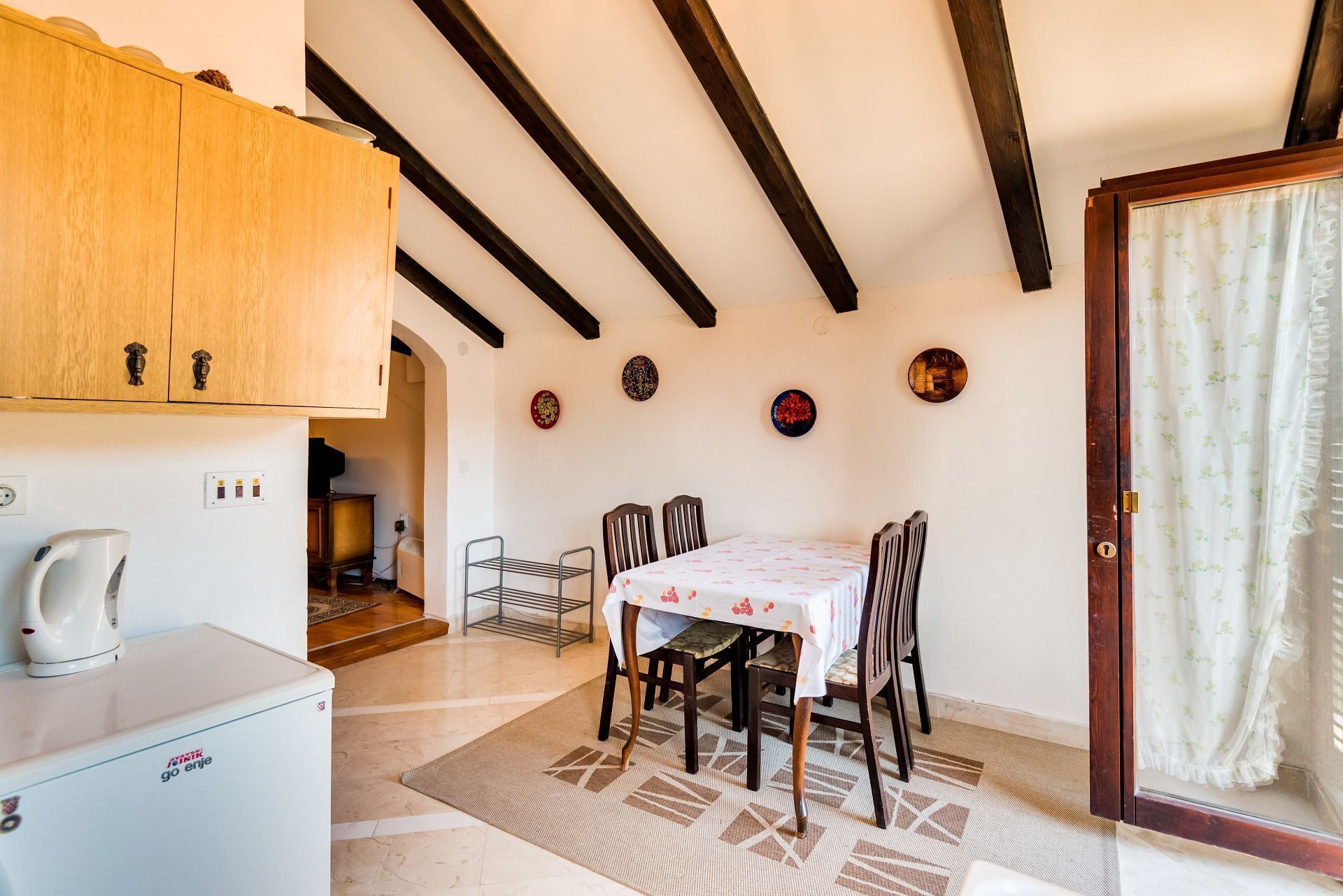 A dining area with a table set for four, wooden cabinets above, a kitchen area with a kettle, and a patterned rug on the floor.