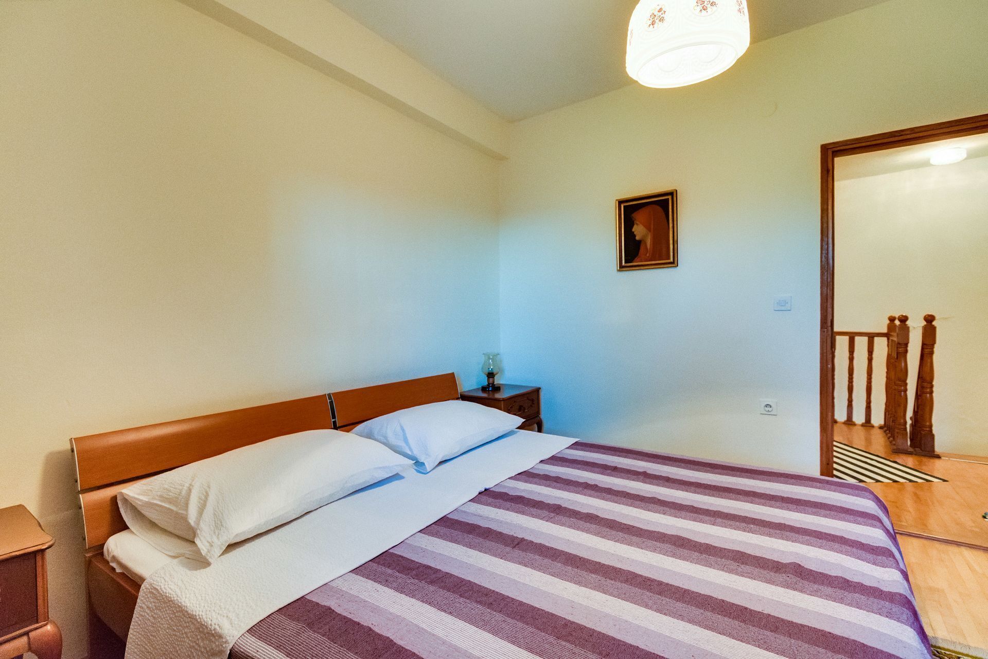 A bedroom with a bed, wooden headboard, a nightstand, a lamp, a framed picture on the wall, and a wooden staircase.