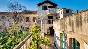 Un edificio tradizionale con un balcone, una scala che conduce al secondo piano e un'area giardino con vegetazione.