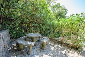 A stone table and bench set in a garden with dense foliage. - Apartments Ani (Crikvenica)