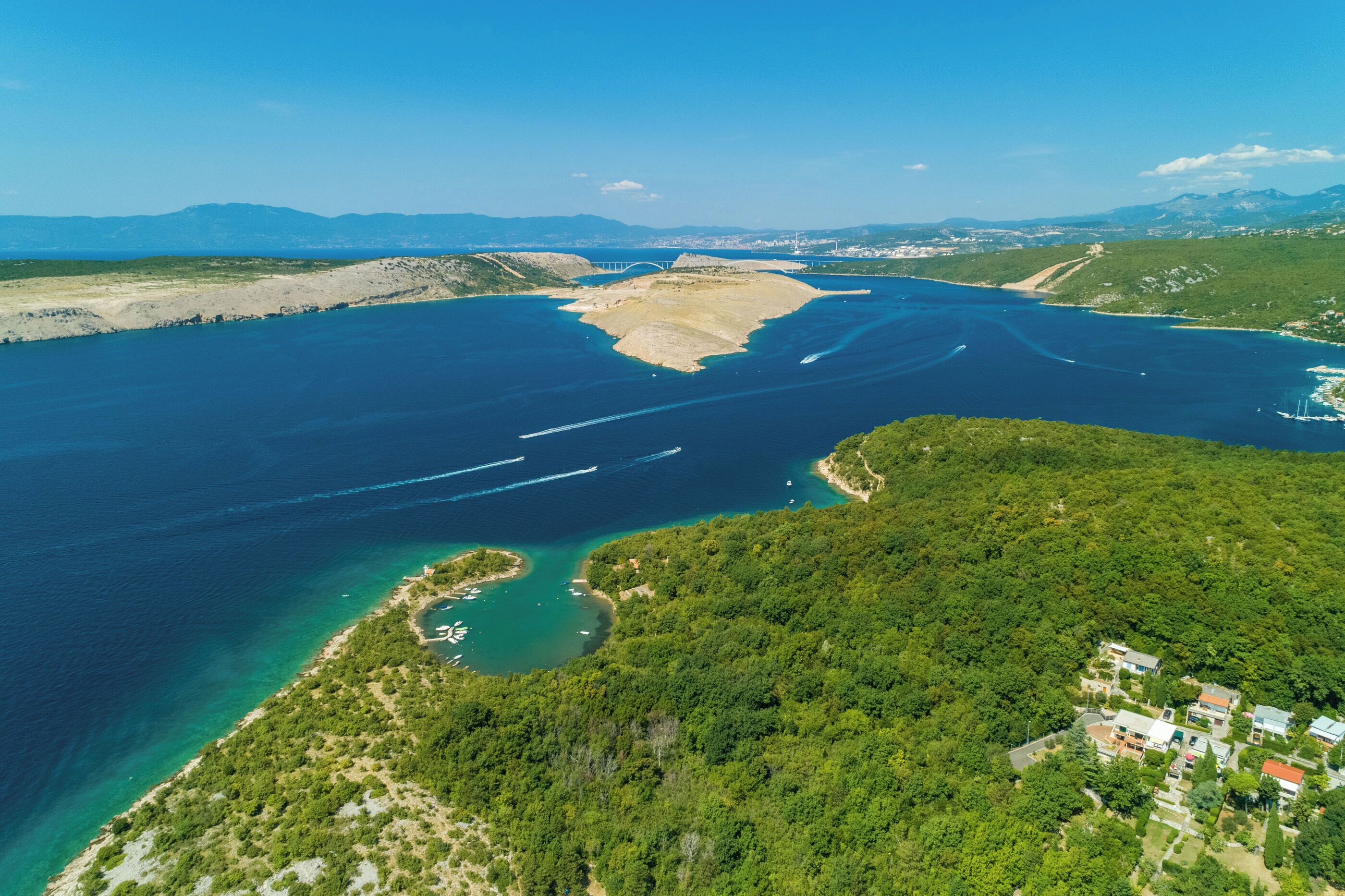 Une vue aérienne d’une zone côtière avec une petite île, des eaux d’un bleu limpide et un pont au loin.