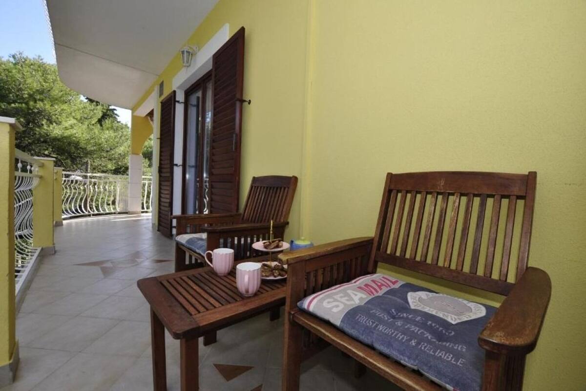 A wooden bench with a cushion, a small table, and a pink mug on a balcony with a yellow wall and a white railing.