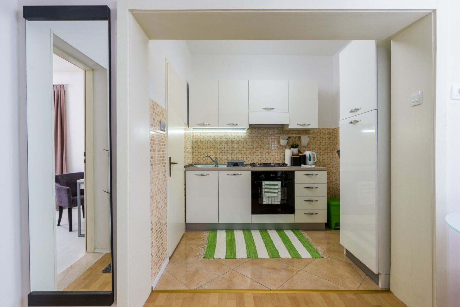 A narrow hallway leading to a kitchen with white cabinets, a tiled backsplash, and a green striped rug.