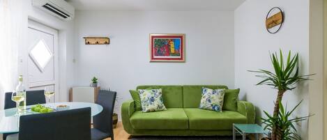 A living room with a green sofa, two side tables, a small potted plant, a framed picture on the wall, and a dining area with a table and chairs.