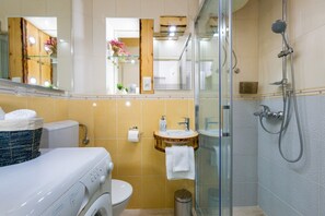 A modern bathroom with a glass-enclosed shower, a white toilet, a washing machine, and a sink with a wooden countertop. - Apartment Jurica (Kastela)