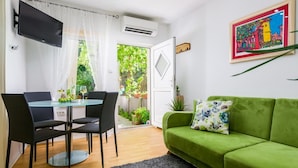 Una sala moderna con un sofá verde, una mesa de comedor con sillas, un televisor de pantalla plana y una puerta que da a un jardín.