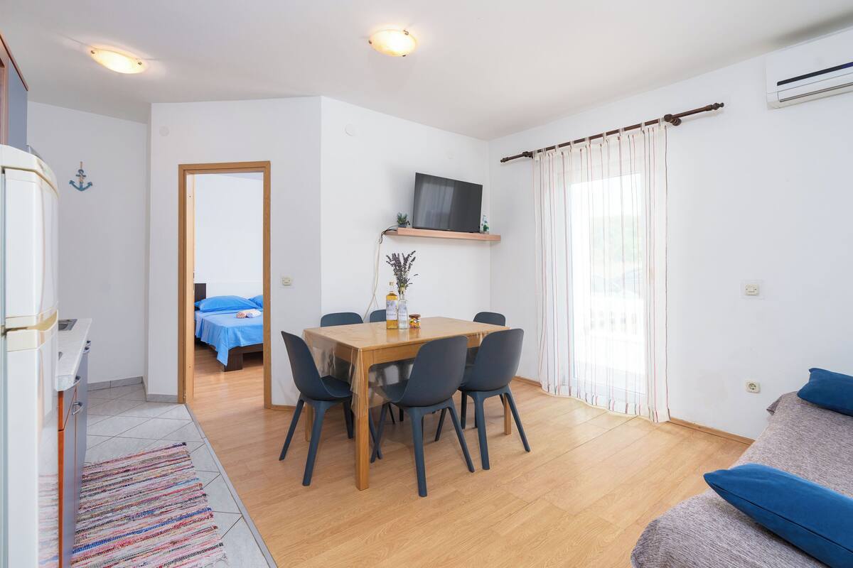 A compact living space with a dining area, a sofa, a TV mounted on the wall, and a bed in the background.