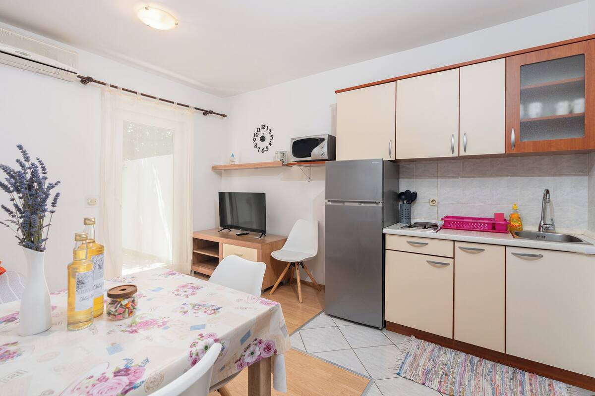 A modern kitchen with a refrigerator, microwave, and wooden cabinets. There is a dining table with chairs and a TV on a stand.