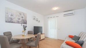 A modern living room with a round wooden dining table, grey upholstered chairs, a flat-screen TV, a sofa with striped cushions, and a large abstract painting on the wall.