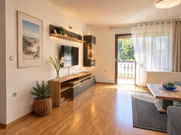 A modern living room with a wooden floor, a flat-screen TV mounted on the wall, a dining area with a table and chairs, and a large window with curtains. - Apartment Ani (Rab)