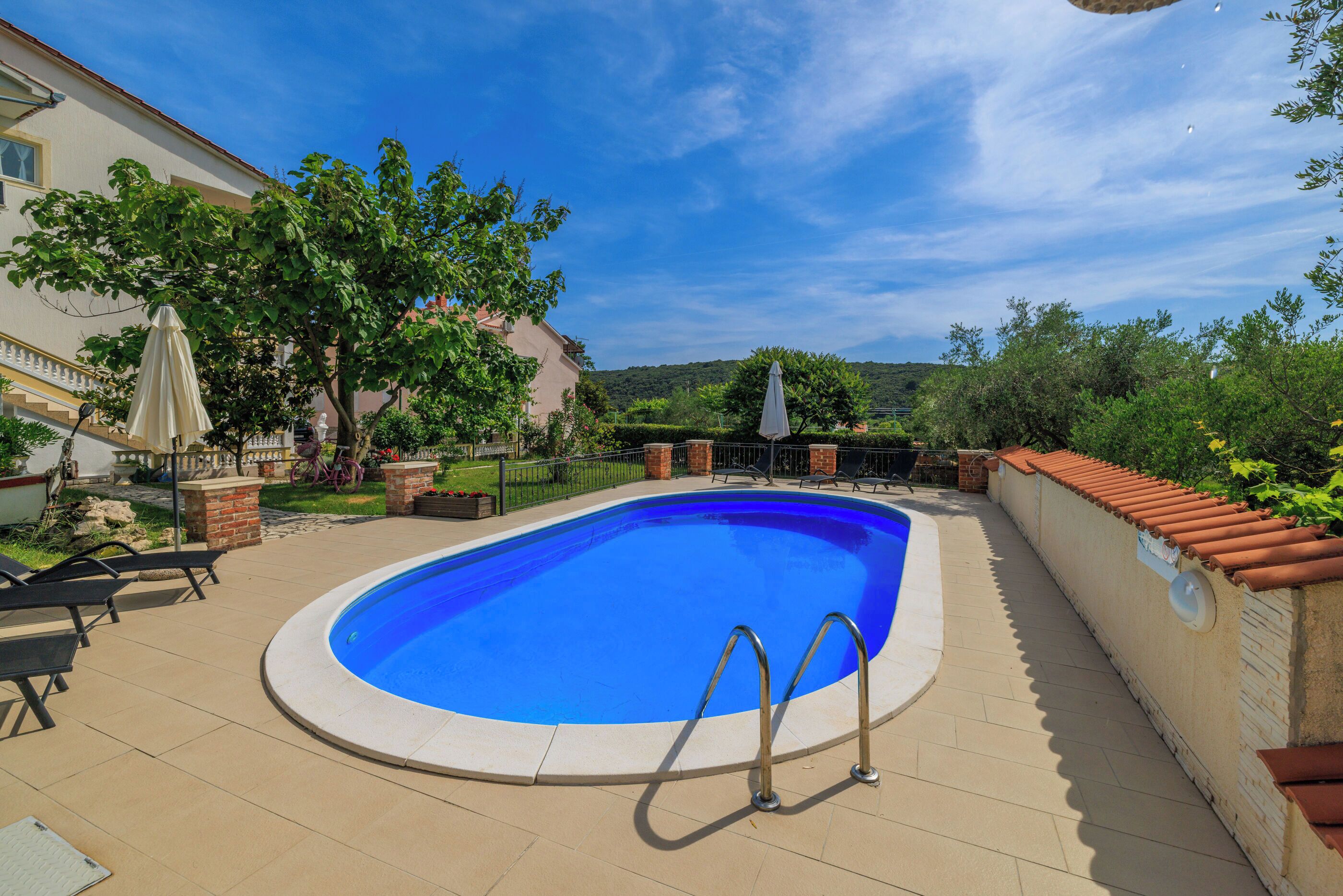 Una piscina con solárium empedrado, rodeada de sillones de descanso y sombrillas, con vistas a una colina al fondo.