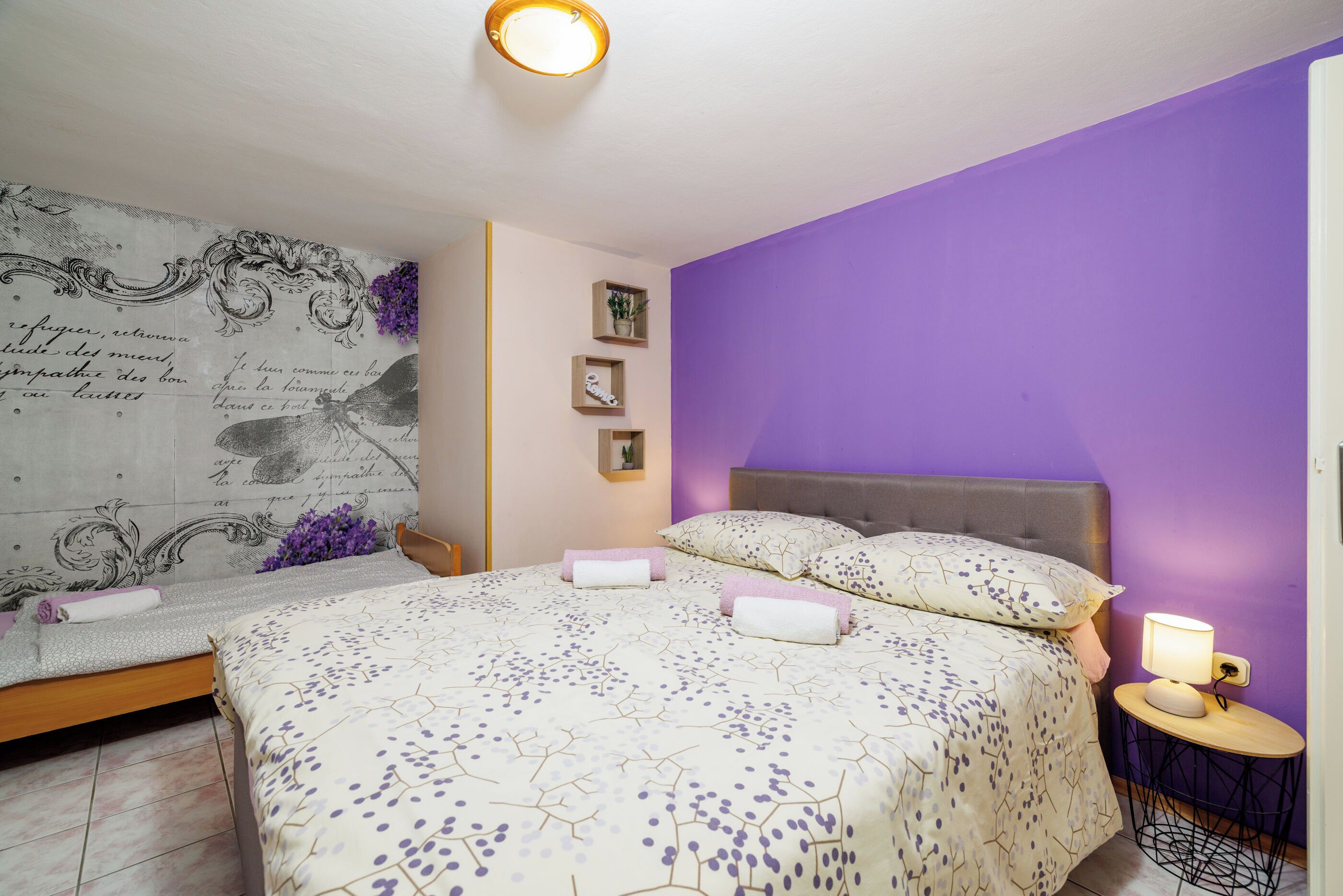 A bedroom with a bed, a bedside table with a lamp, a wall with decorative elements, and a small shelf with items.