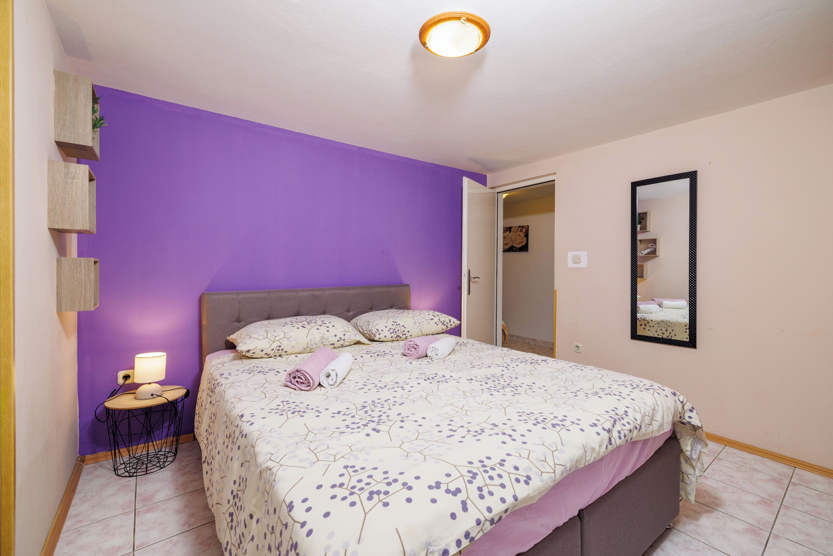 A bedroom with a purple wall, a bed with a floral bedspread, a bedside lamp, and a mirror on the wall.