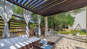 An outdoor patio with wicker furniture, a stone floor, and a pergola with white curtains. - Apartments Roby (Rab)