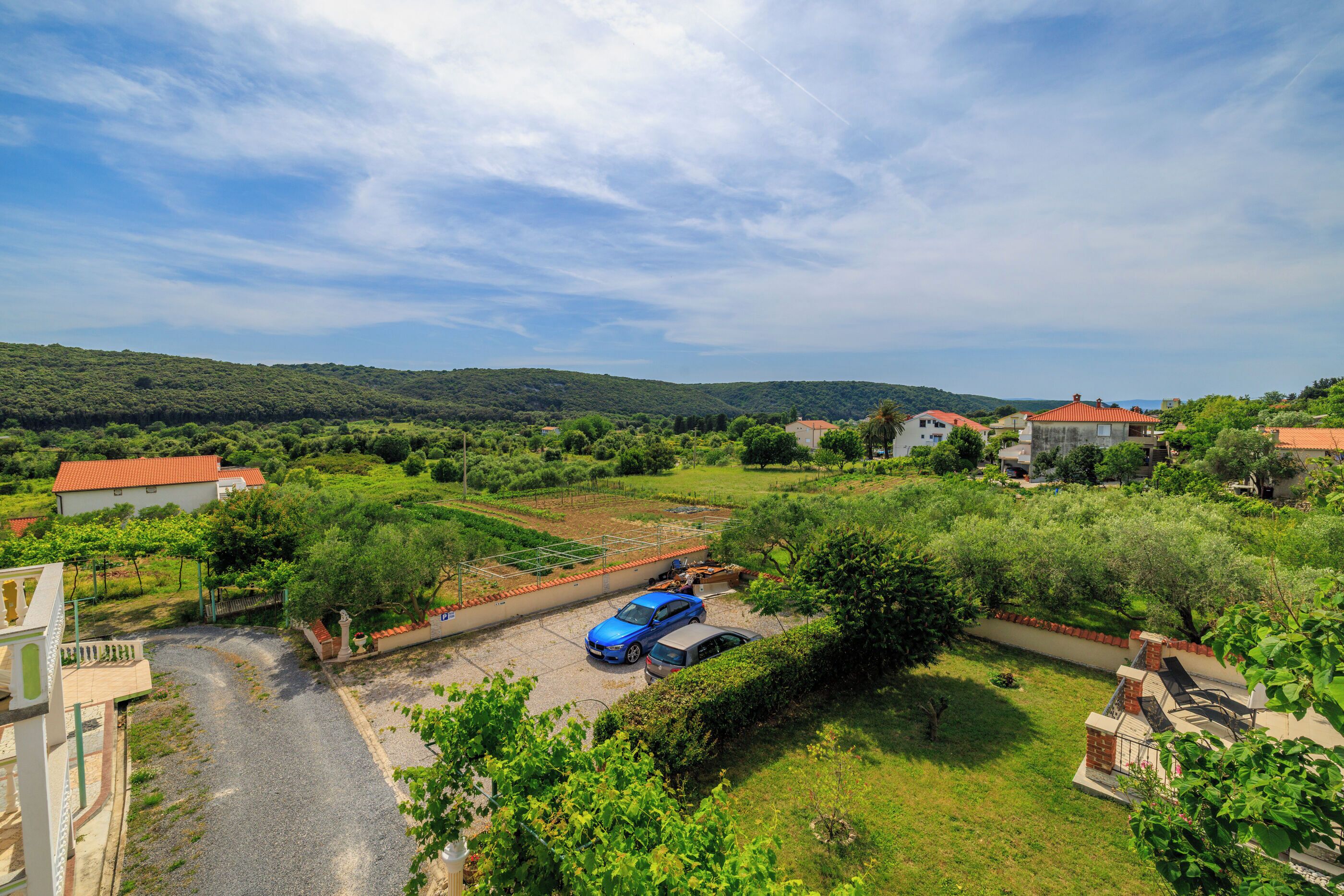 A rural landscape with a paved driveway, parked cars, and houses with red roofs.