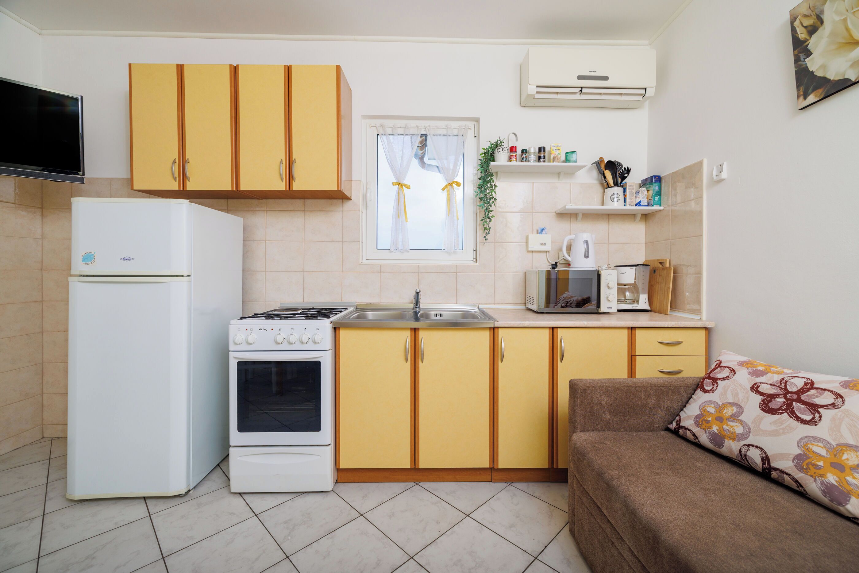 Une cuisine compacte équipée d’un réfrigérateur, d’une cuisinière, d’un four et de placards jaunes. On y trouve une fenêtre avec des rideaux, un évier et un four à micro-ondes. Un canapé marron avec un coussin à motifs floraux se trouve dans un coin.