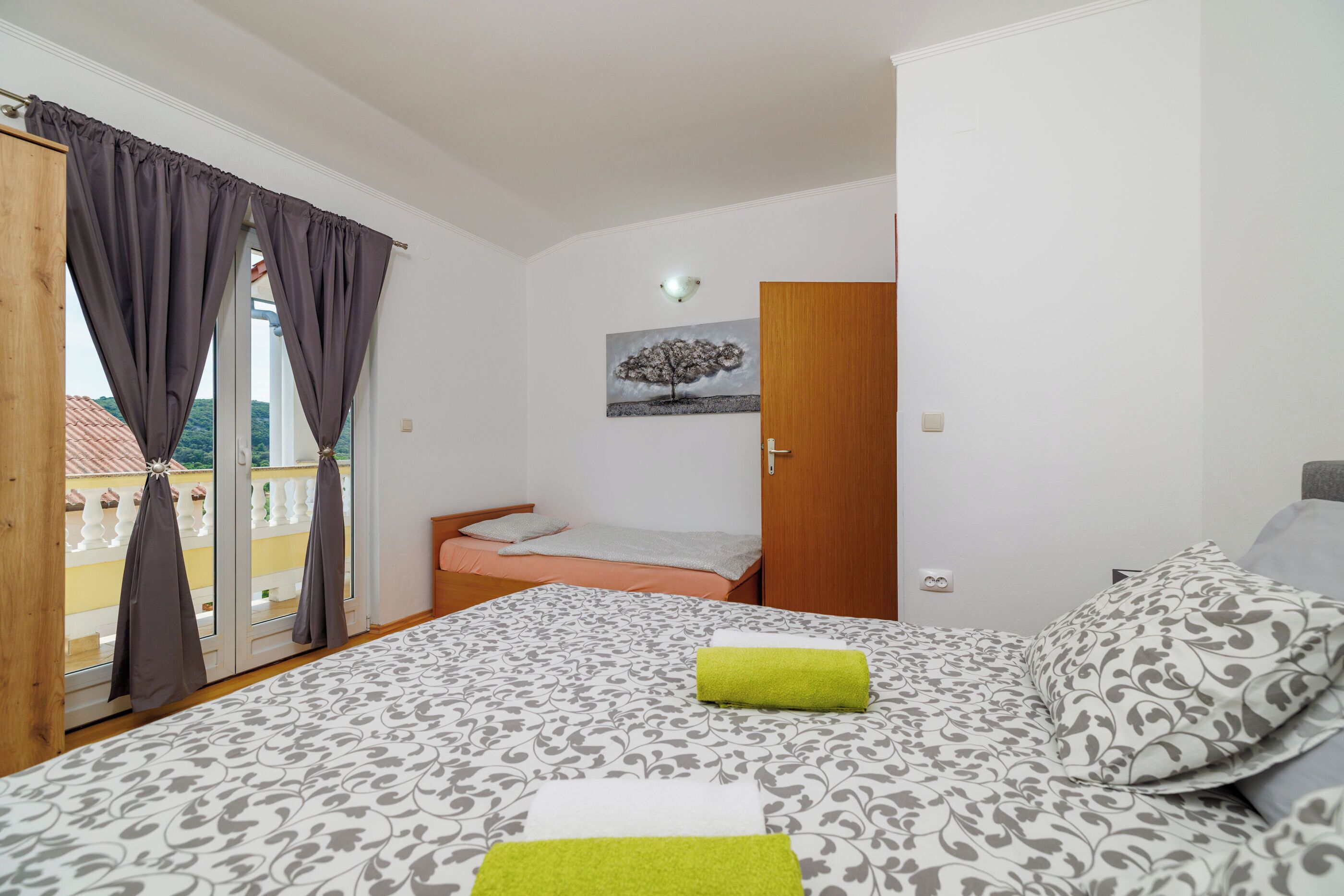 A bedroom with a bed, a window with curtains, a painting on the wall, and a balcony visible through the window.