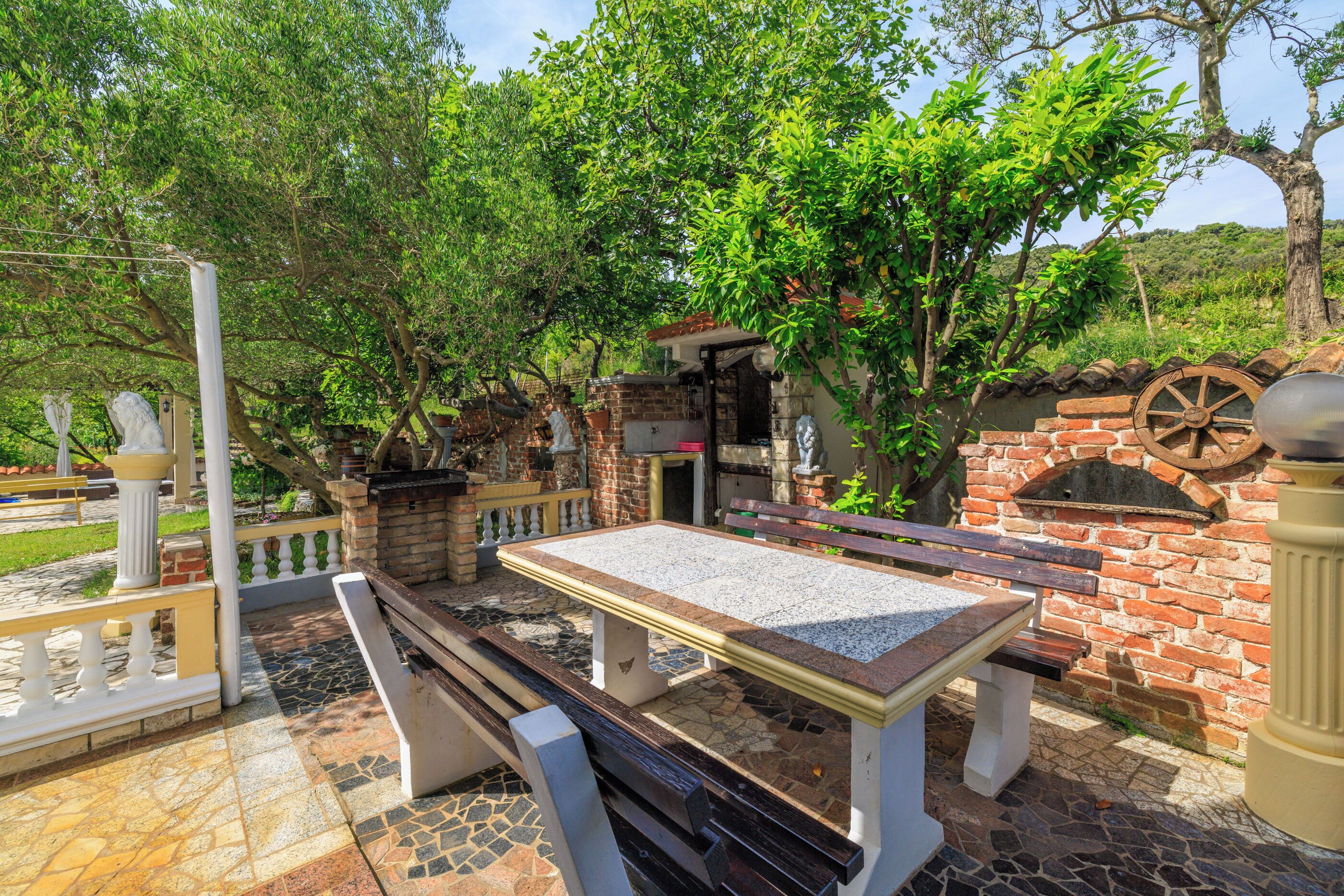 Eine Terrasse im Freien mit einem Steintisch und Bänken, umgeben von Ziegelmauern und Grünpflanzen.