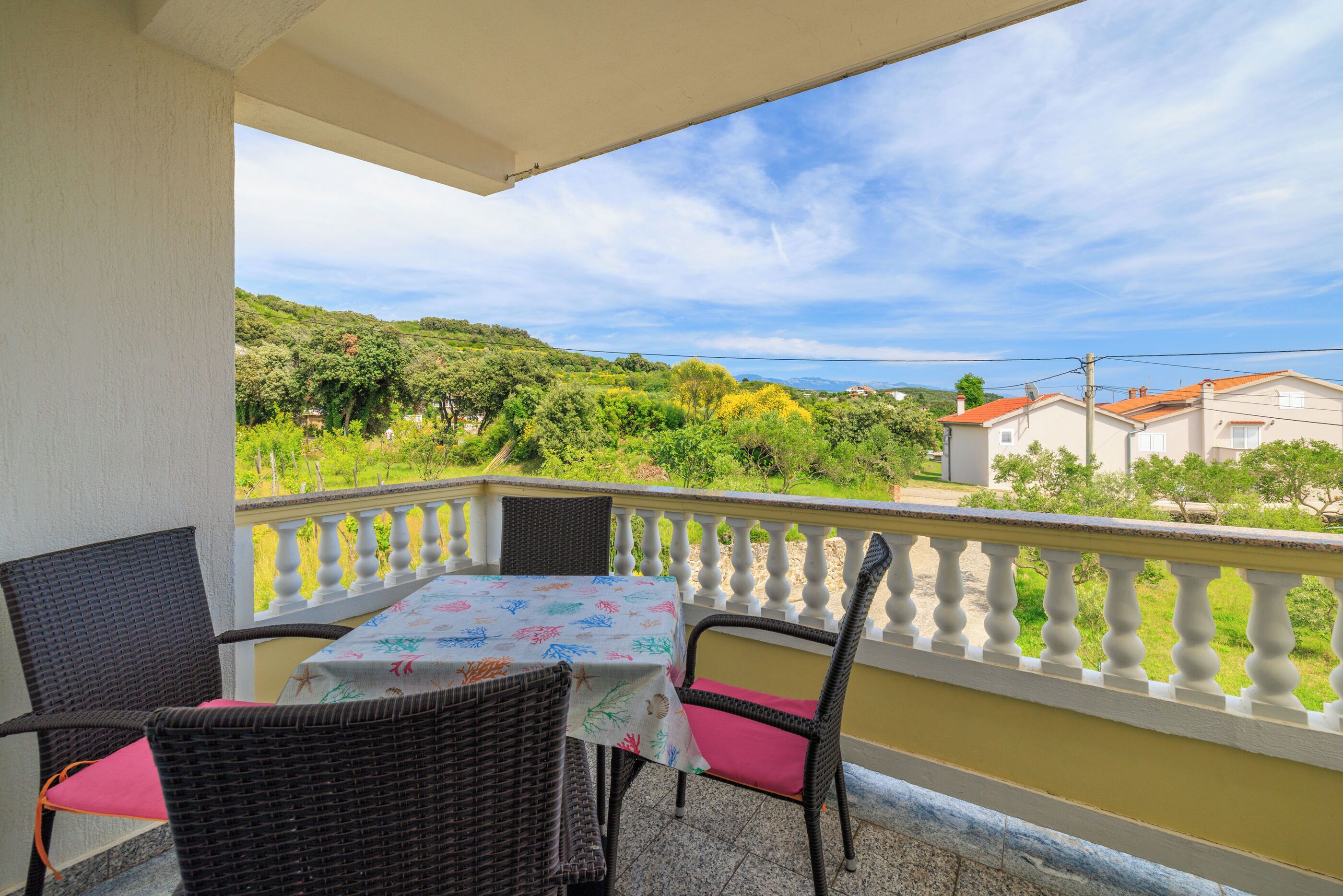 A balcony with wicker chairs and a table, overlooking a green landscape and buildings.