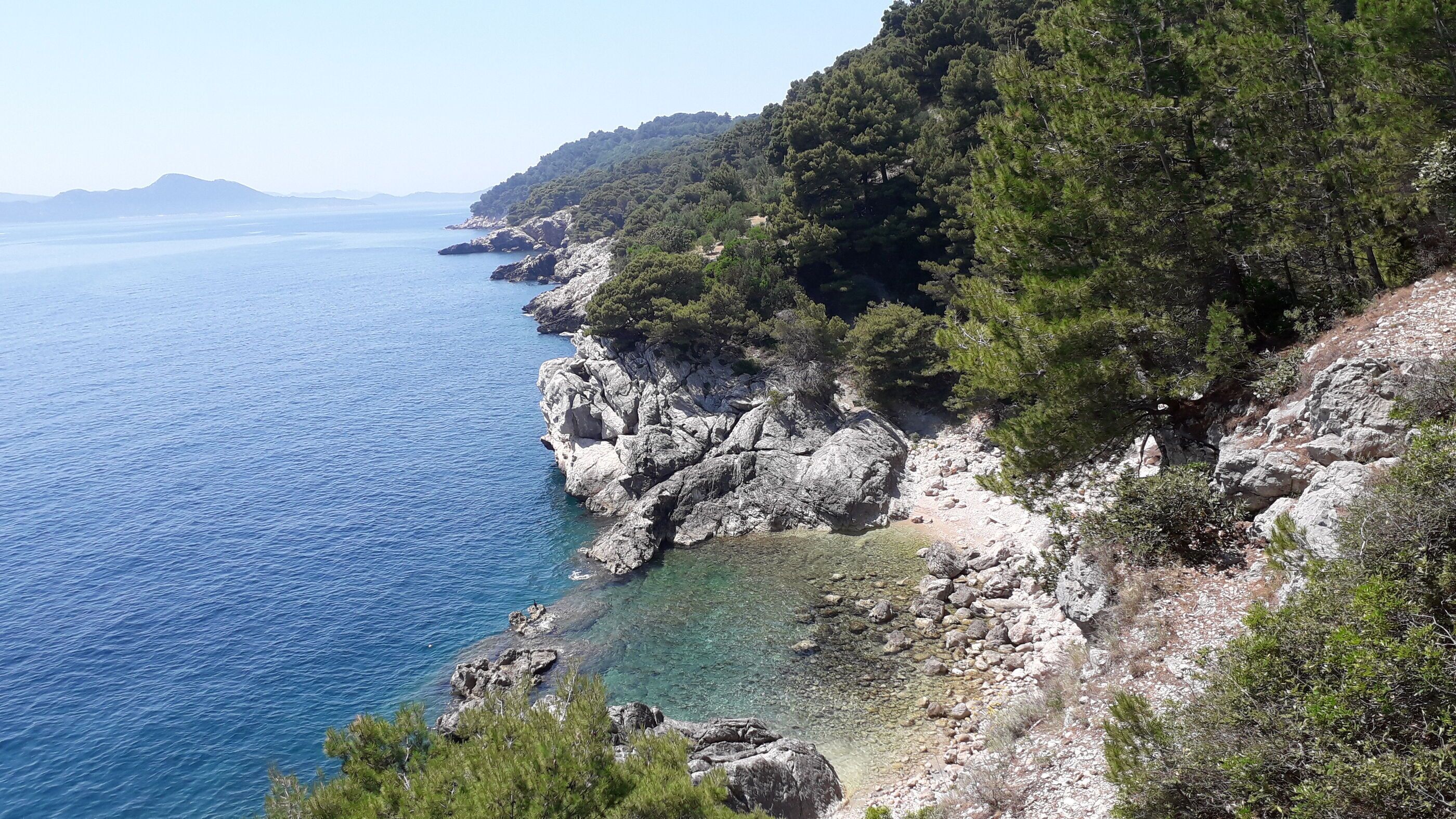 A coastal landscape with rocky cliffs, clear blue sea, and dense greenery.