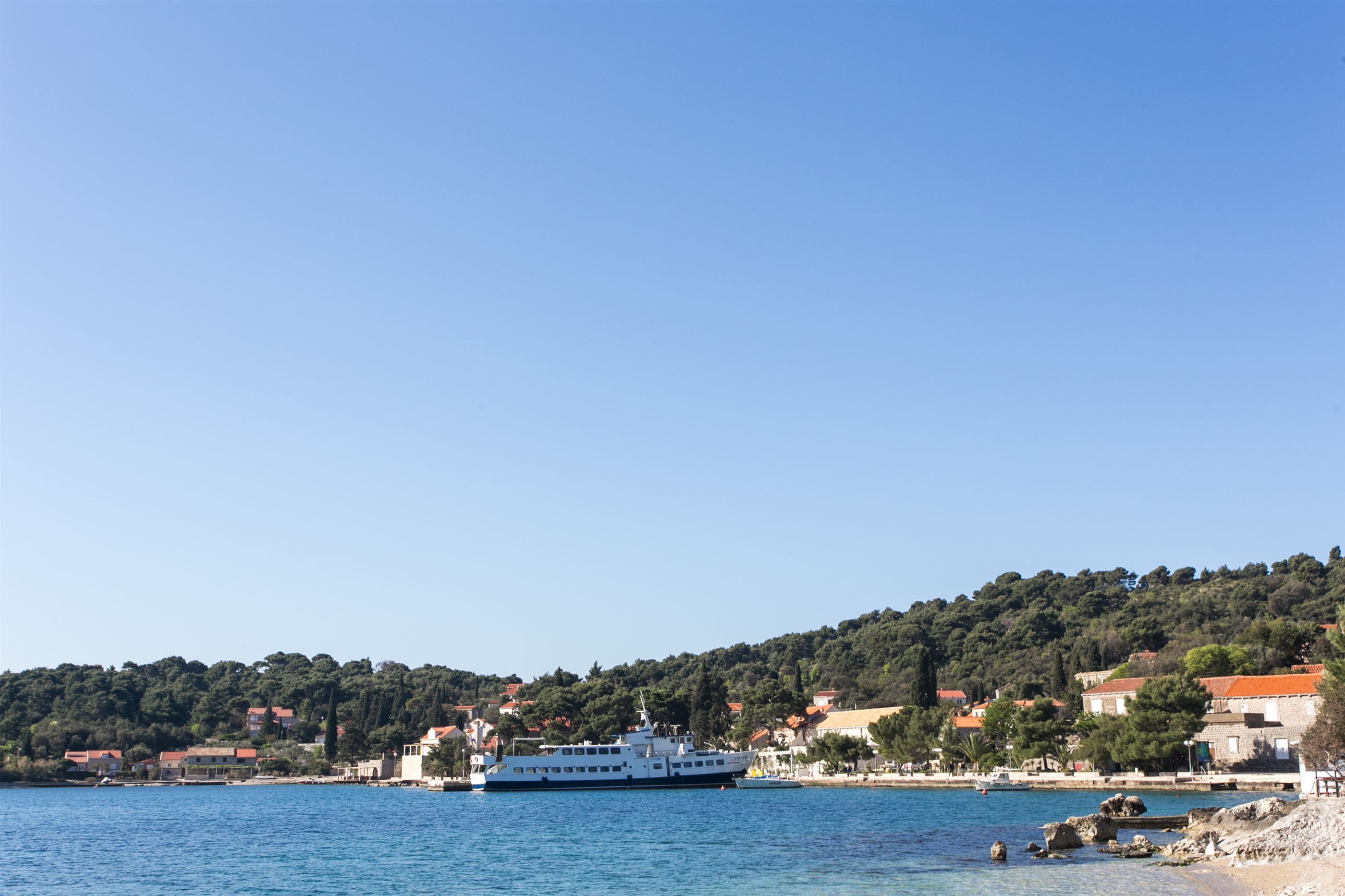 A coastal town with a large white boat docked near the shore, surrounded by buildings with red roofs and lush green hills in the background.