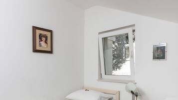 A single-bed room with a window, a bedside table with a lamp, and two framed pictures on the wall.
