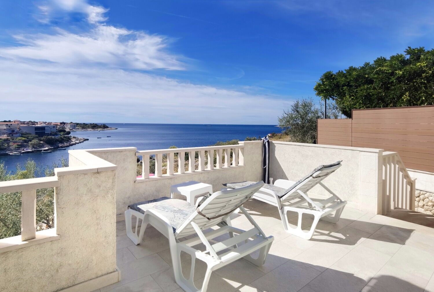 A terrace with white plastic furniture, a view of the sea, and a distant town.