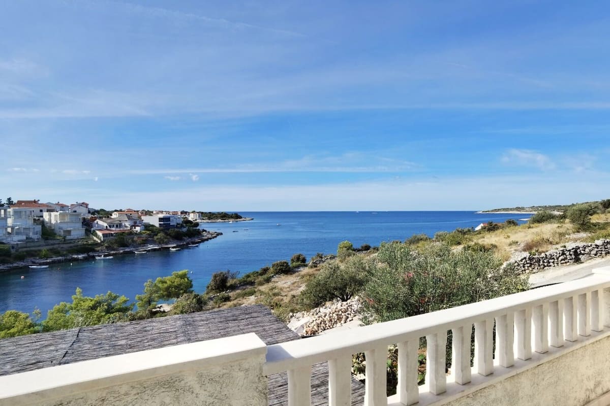 Un pueblo costero con edificios a lo largo del litoral, un cielo azul despejado y una ladera rocosa con vegetación escasa.