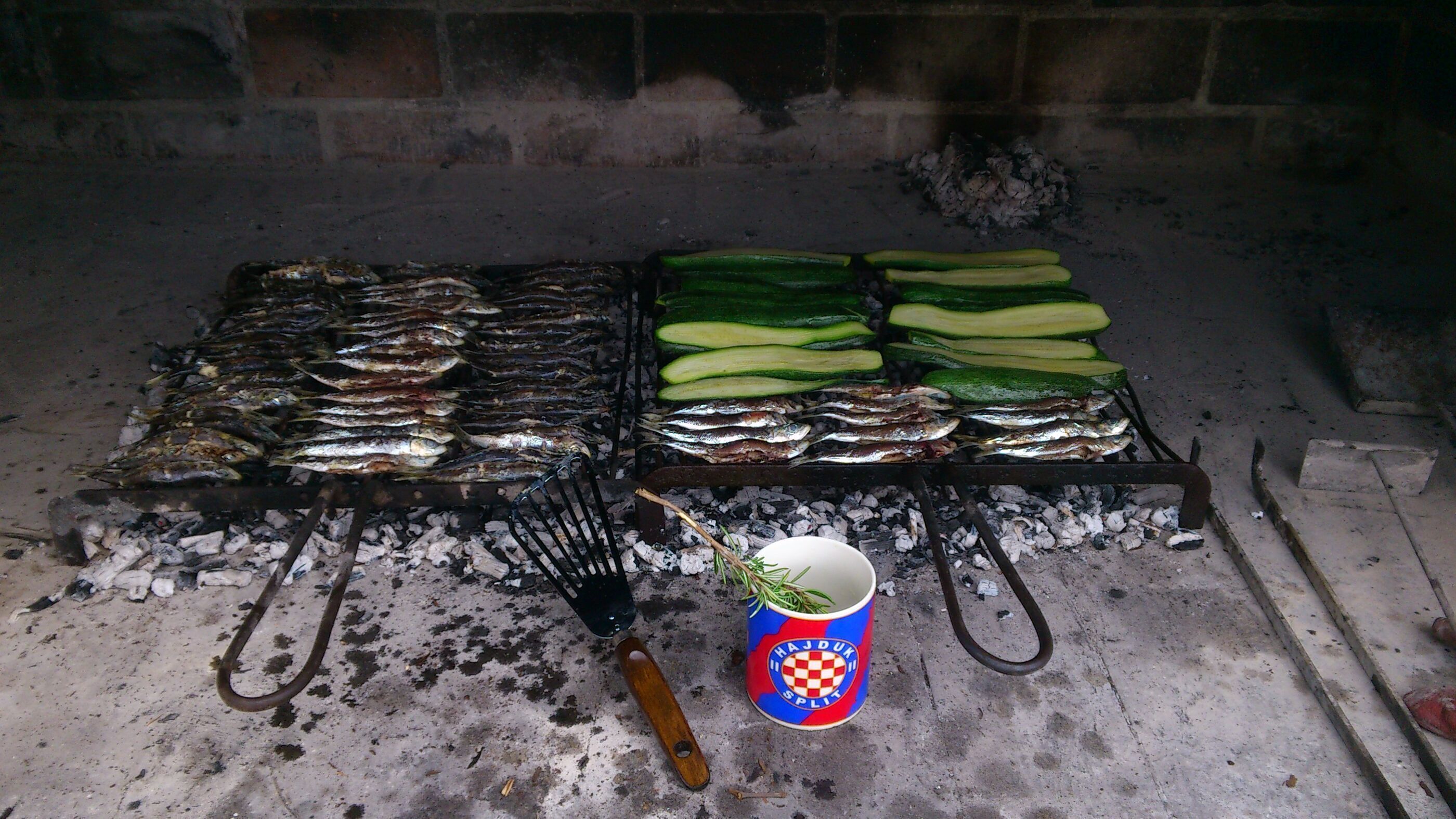 Un gril avec du poisson et des légumes, une fourchette en métal et une tasse à l’effigie d’une marque, le tout posé sur une surface en béton.