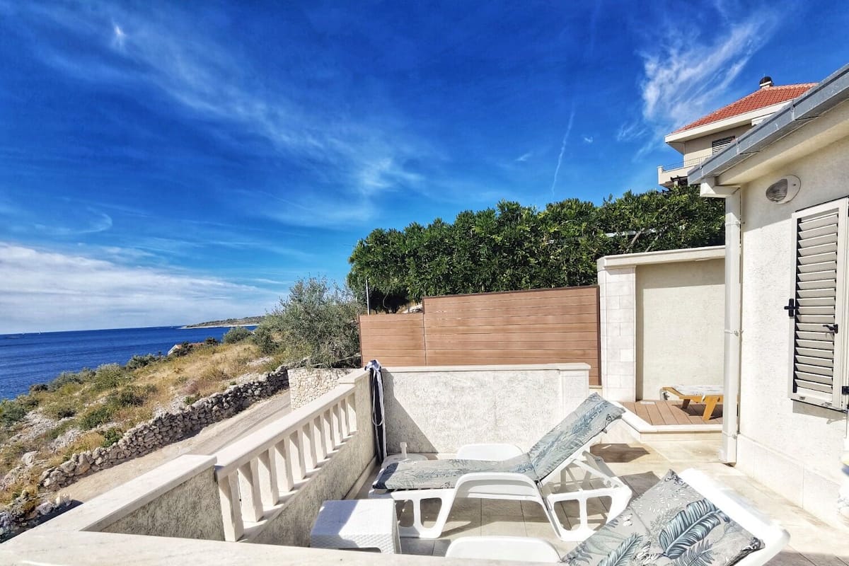 Un patio con sillones de descanso y una mesa, con vistas a una zona costera con valla de madera y muro de piedra.