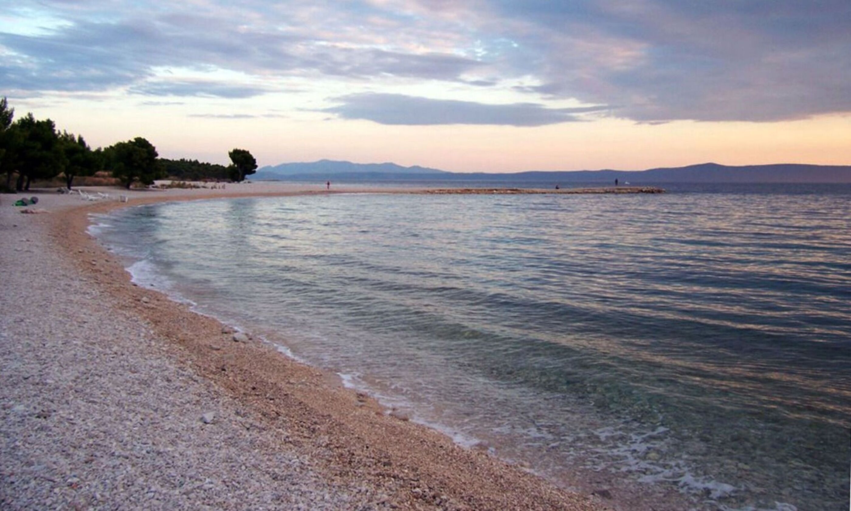 Ein Strand mit klarem Wasser, Kieselsteinen und fernen Bergen.