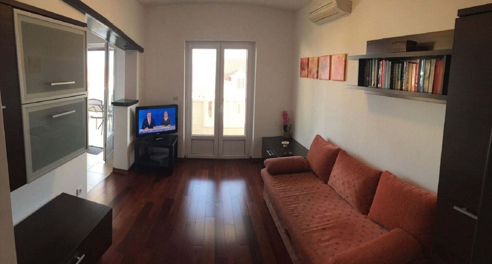 A living room with a wooden floor, a sofa, a television, a bookshelf, and a door leading outside.