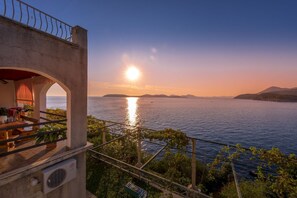 Un balcon offrant une vue sur la mer au coucher du soleil, une terrasse avec une table et des chaises, et un bâtiment doté d’une façade en pierre.
