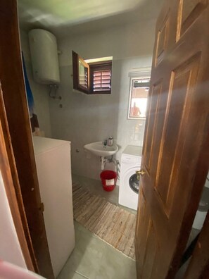 A compact bathroom with a sink, a window with blinds, a washing machine, and a red bucket on the floor. - Apartment Jakov (Okrug)