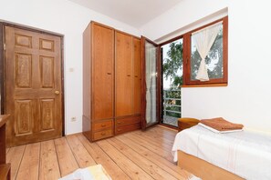 A room with a wooden door, a wardrobe, a bed with a white bedspread, and a window with a view of greenery. - Apartment Jakov (Okrug)
