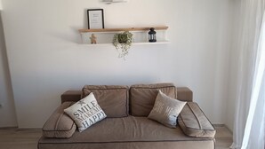 A modern living room with a beige sofa, two pillows with motivational text, a white wall with a framed picture, and a wooden shelf with decorative items. - Apartments Antonia (Okrug)