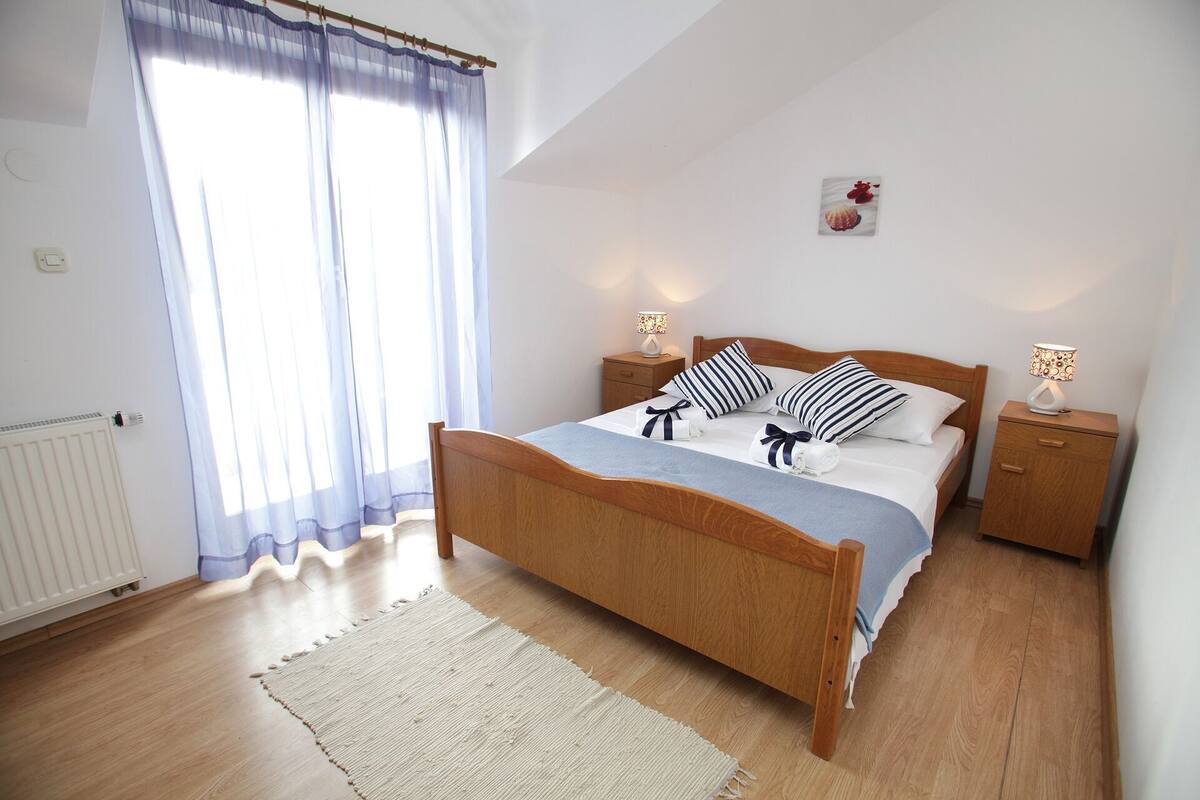 A bedroom with a wooden bed, two bedside tables with lamps, a radiator, and a window with sheer curtains.