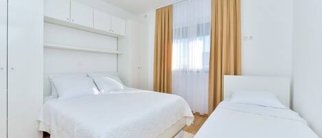 Une chambre avec deux lits, du linge de lit blanc, un plancher en bois et une grande fenêtre avec des rideaux.