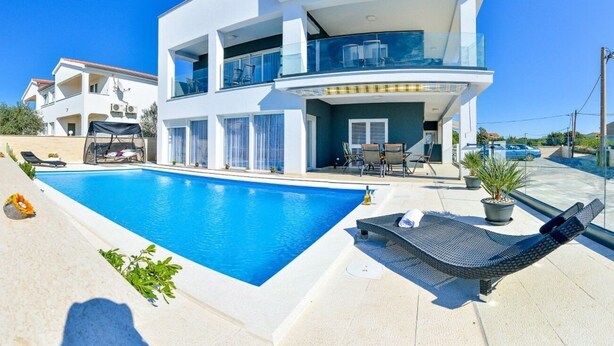 A modern two-story building with a swimming pool, outdoor seating, and a clear blue sky. - Apartments Sunny by the Sea (Nin)