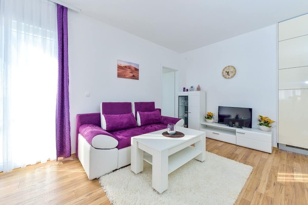 A modern living room with a purple sofa, white coffee table, and a TV on a white cabinet.