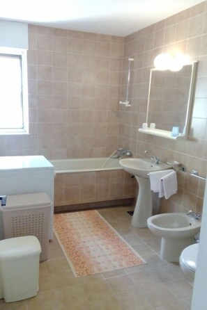 A bathroom with a bathtub, sink, and bidet. There is a window, a towel rack, and a patterned rug. - House Marus (Omis)